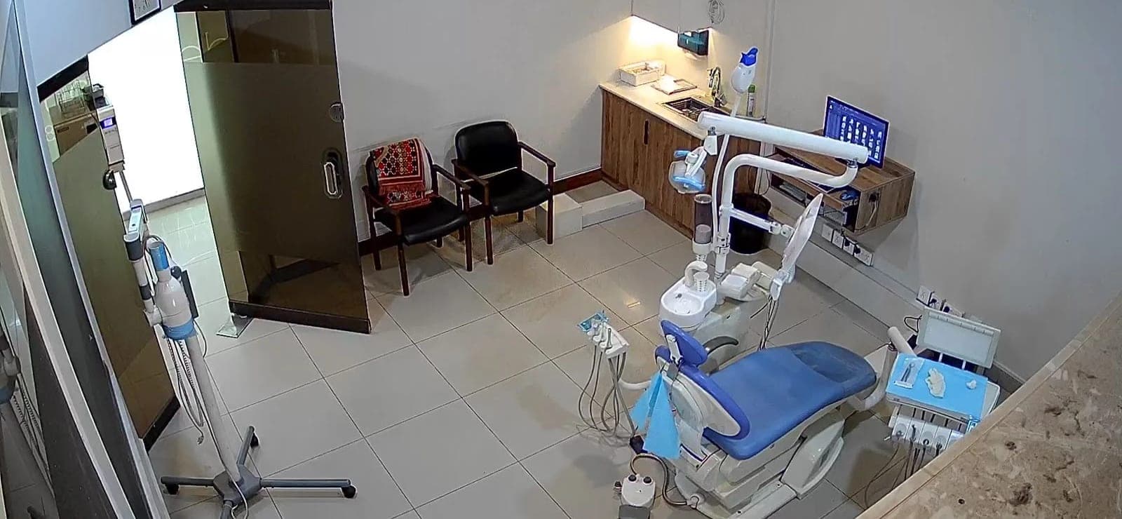 A second treatment room at Dental Plus: a blue dental chair, overhead light, a wooden counter with a sink, a monitor and two chairs